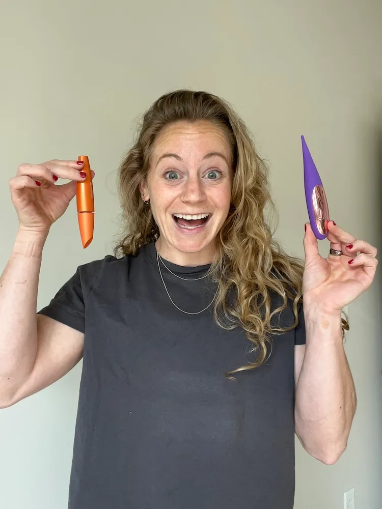 Blonde woman in black t-shirt with two necklaces holding orange mascara tube in one hand and purple vibrator in another