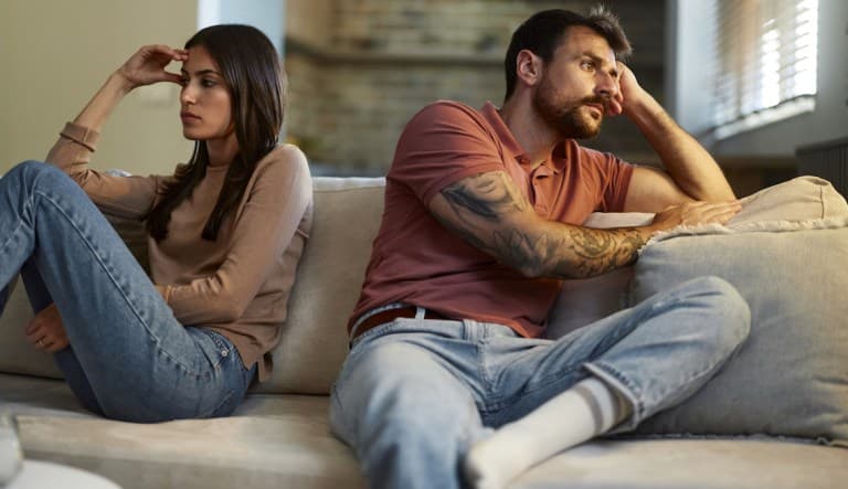 Worried couple sitting back-to-back on sofa at home while having problems in their relationship. This photo is being used to promote an article about the different types of manipulation