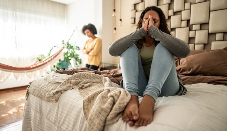 A worried young woman is sitting on her bed at home and covering her face. In the background, another woman is leaning against the wall with her arms crossed, looking equally upset. This photo is being used in an article about relationships fizzling out.