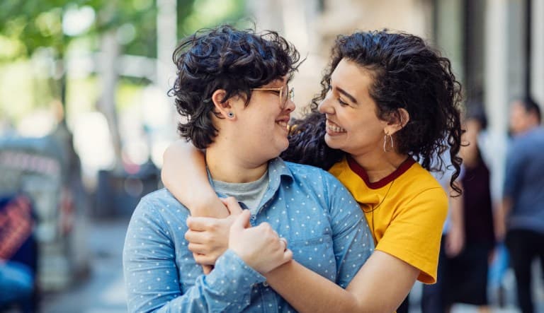 Two women embrace on the sidewalk on a sunny day. They are smiling at each other and appear to be in a relationship. This photo is being used to promote an article answering the question