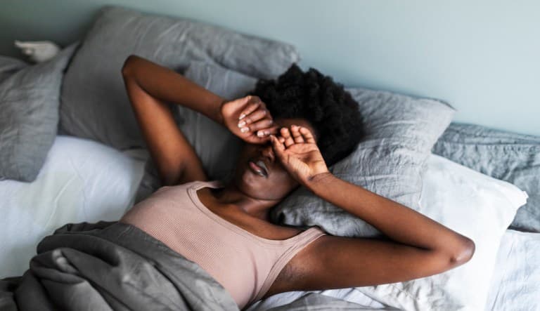A young Black woman yawns and rubs her eyes in bed. Her hands are resting over her face and she is lying against two gray pillows with a blanket covering her torso. This photo is being used in an article answering the question