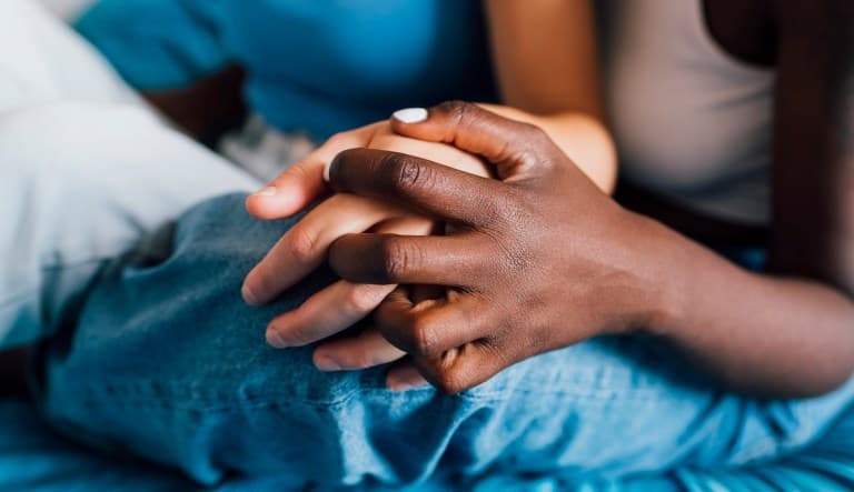 A close-up shot of two people holding hands. One person has dark skin and one person has light skin and their clasped hands are resting on a jean-clad knee. This photo is being used to promote an article answering the question