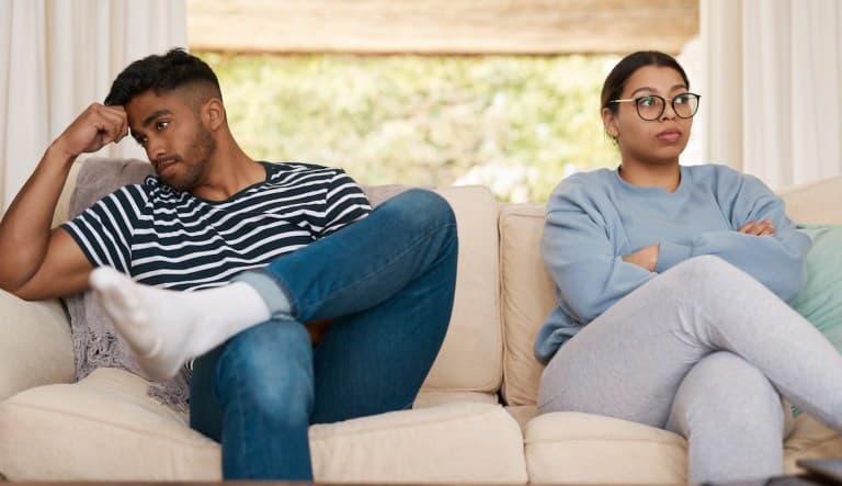 A man and woman ignore each other, facing different directions while sitting at home on the couch. The woman