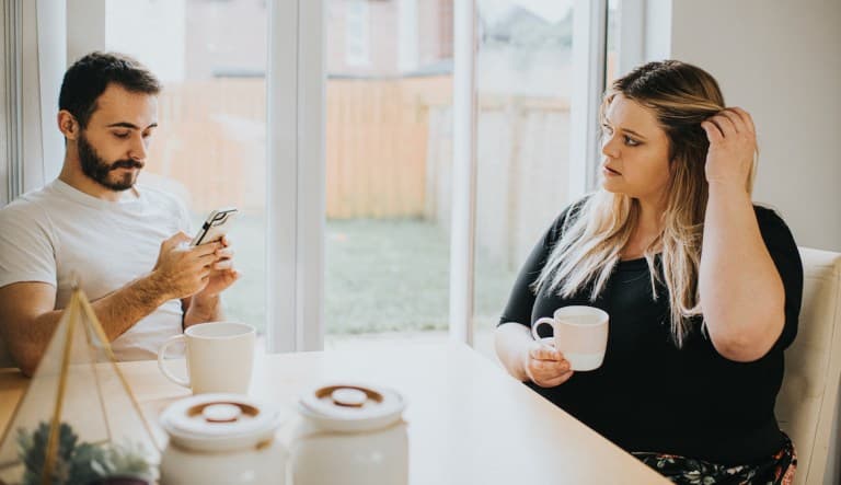 A woman and man sit at a kitchen table. The man looks down at his phone and subsequently ignores the woman. The woman looks insecure and hurt as she looks at him, hoping for some attention. This photo is being used in an article about signs of an untrustw