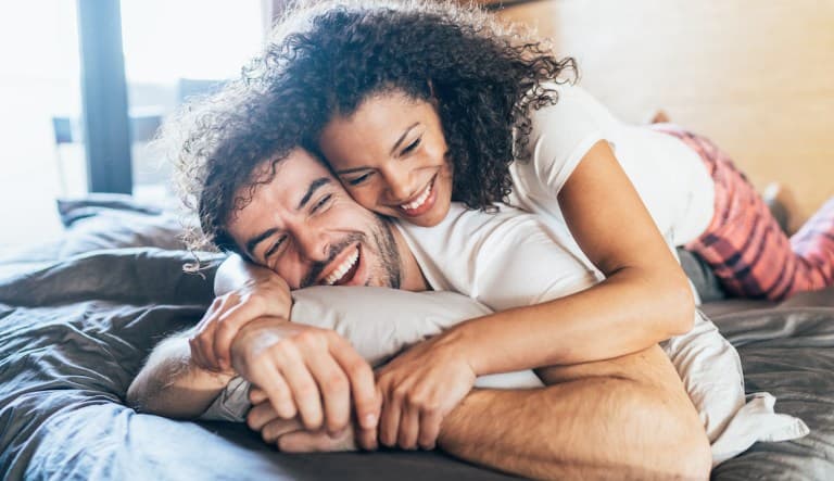 A beautiful young couple enjoys leisure time in bed. The woman has curly black hair and is laying on top of the man, who is laying on his stomach. They are both smiling and have their arms interlinked. This photo is being used in an article about sex pref