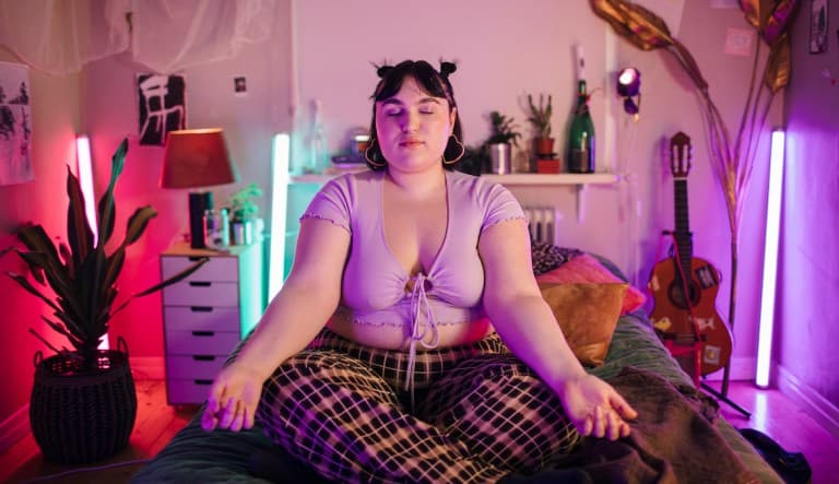 Young woman meditating on a bed in a colorfully-illuminated bedroom. This photo is being used to promote an article about anxiety crying.