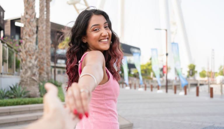 A young woman in a sleeveless pink dress looks back and smiles as she pulls on her boyfriend