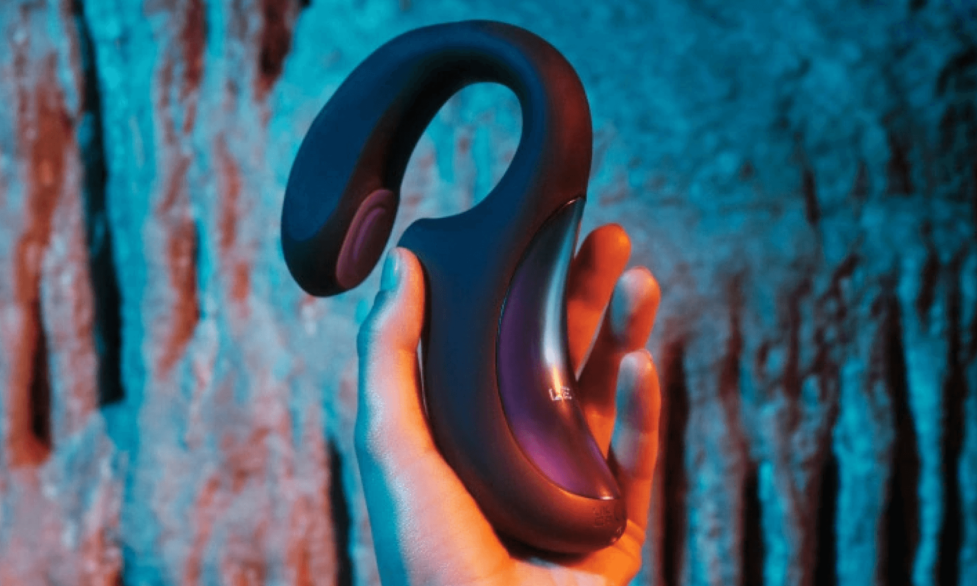 Hand holding black silicon vibrator with iridescent metal accents against cave wall with teal lighting