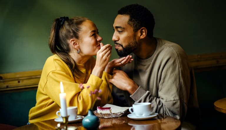 Girlfriend feeding bite of raspberry cheesecake to her boyfriend at coffee shop. Both people are smiling and making eye contact. This photo is being used to promote an article answering the question,