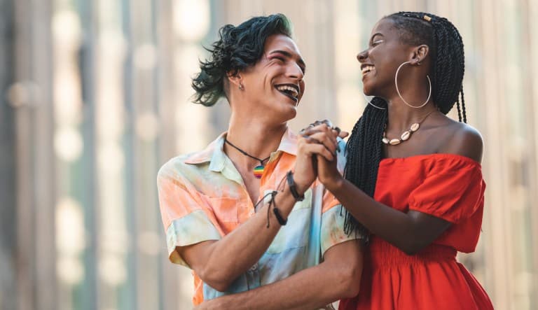 Two people smile and hold hands in a display of friendship. The person on the left has an asymmetrical haircut, dark lipstick, and a multi-colored button-up shirt. The person on the right is wearing a vibrant red dress, a shell necklace, and oversize hoop