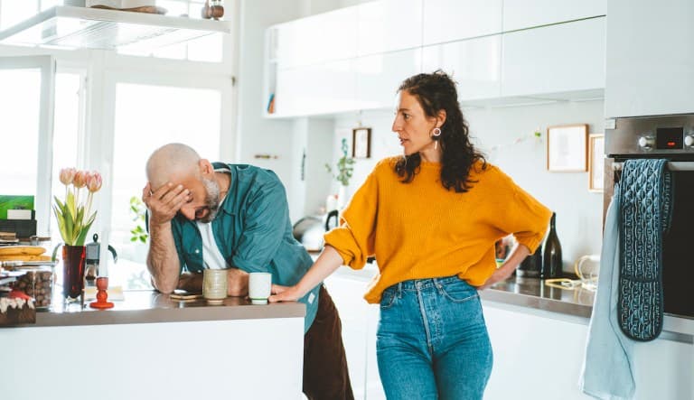 A couple stand in their kitchen looking visibly frustrated. The man is leaning against the island with his face in his hand, and the woman has a hand on her hip as she talks to the man. This photo is being used in an article about the anxious-avoidant dat