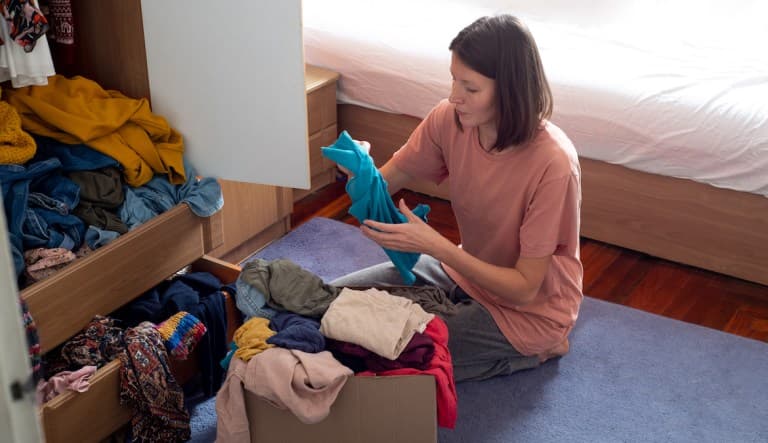 A woman with a lob haircut sits on the ground of her bedroom sorting through her closet. In her hand, she appears to be holding a folded blue t-shirt. In front of her are drawers and boxes full of clothes. This photo is being used in an article about what