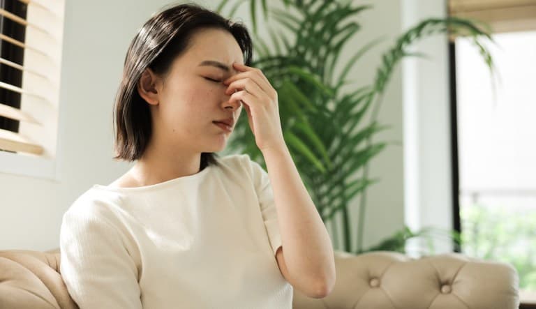 A young woman sits at home on the couch clutching the bridge of her nose as she deals with a headache. This photo is being used in an article about how to erase a bad memory.