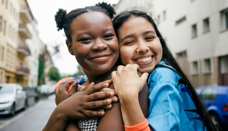 Two women in a loving embrace stand in the middle of the street. One of the women is hugging the other woman from behind and they are both smiling directly at the camera.
