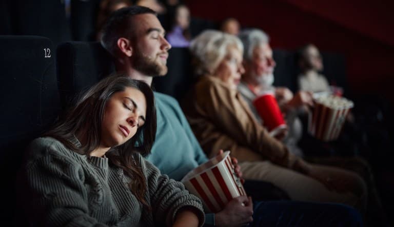 A couple sit in a darkened movie theater surrounded by other moviegoers. The woman is leaning against the man