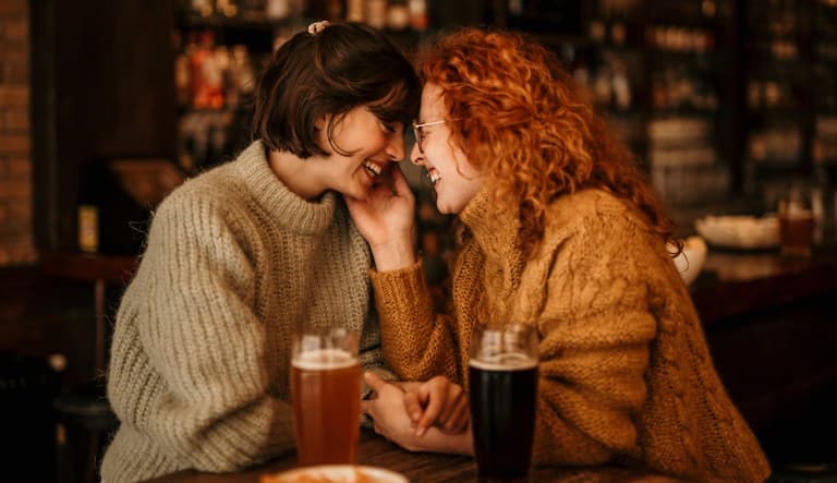 Two women sit in a bar, laughing with their noses almost pressed against each other. One woman is caressing the other woman
